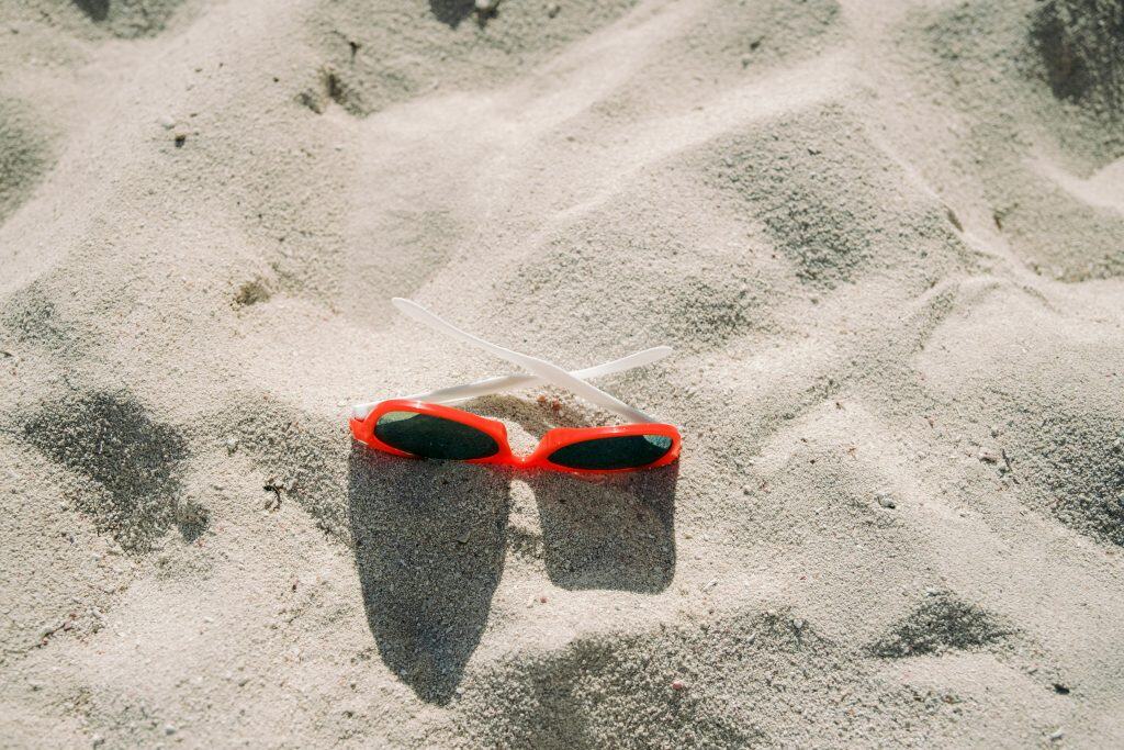 Red sunglasses with white arms lying on light-colored sand, casting small shadows.
